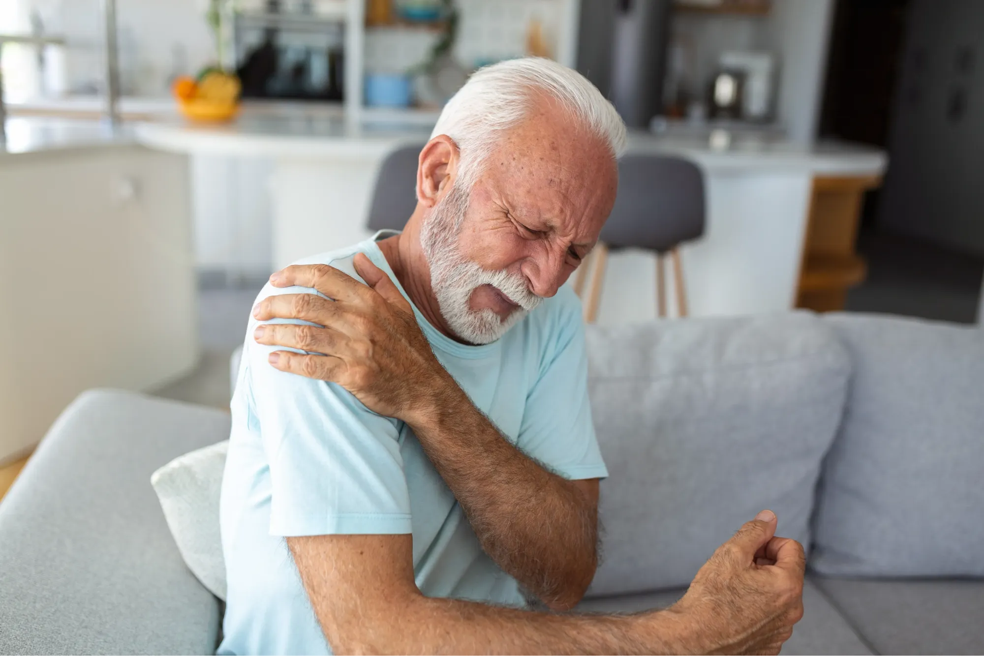 Senior man sitting on a couch at home, wincing and holding his shoulder in pain.